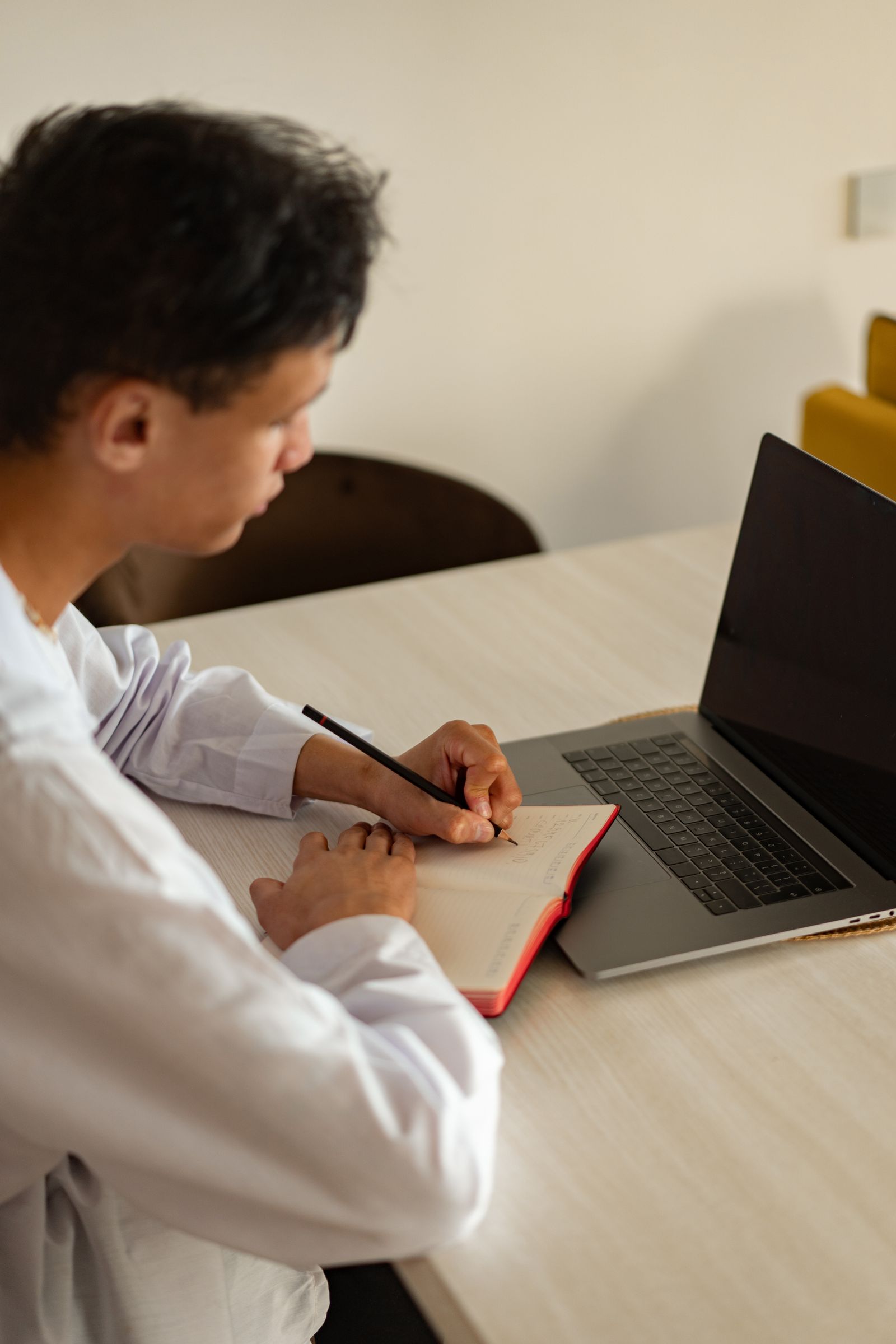 Student writing in a notebook beside a laptop at home